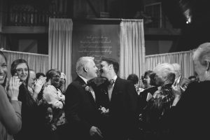 Black and white photograph of LGBT wedding with two grooms about to kiss after their ceremony and stopping in the aisle