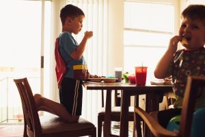 Young boy dressed like Superman eating breakfast at the kitchen table with his little brother