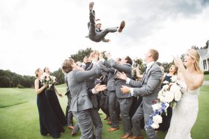 Groom being thrown into the sky above groomsmen while bride and bridesmaids watch in surprise.