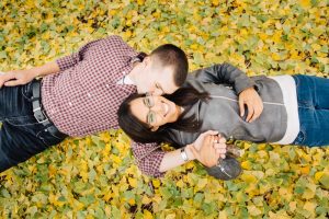 Inter-racial couple holding hands and smooching while laying in a pile of yellow gingko leaves
