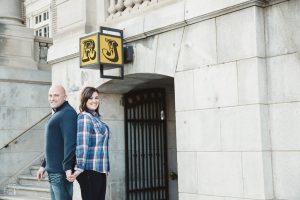 Engaged couple standing back to back and holding hands in front of stone building