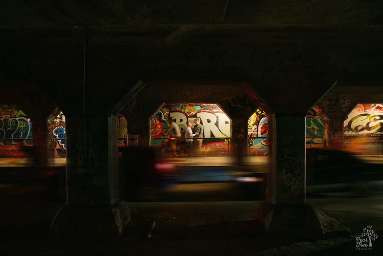 Two men standing close in Krog Street Tunnel as cars and bicyclists move by in a blur.