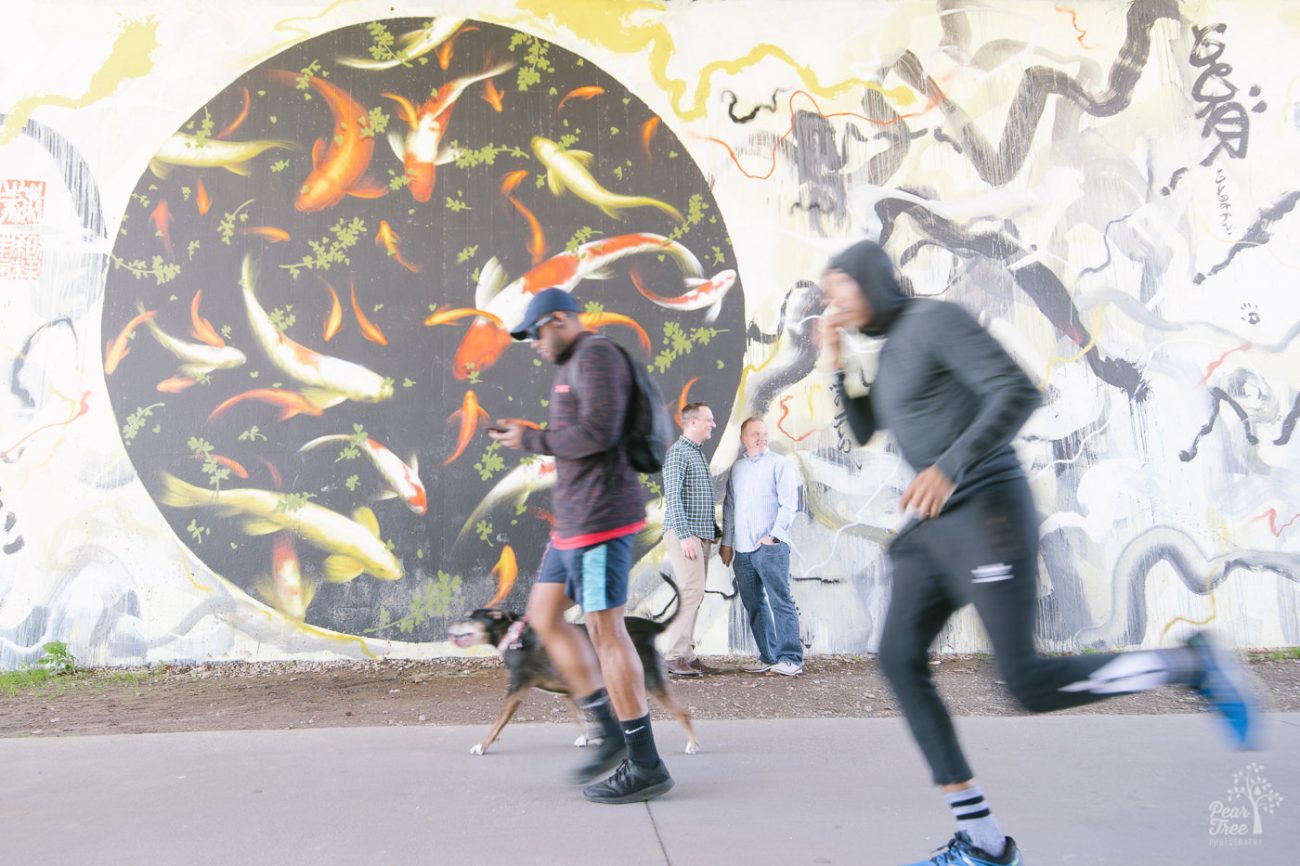 Two men standing in front of koi mural along Atlanta Beltline while joggers run past in a blur.