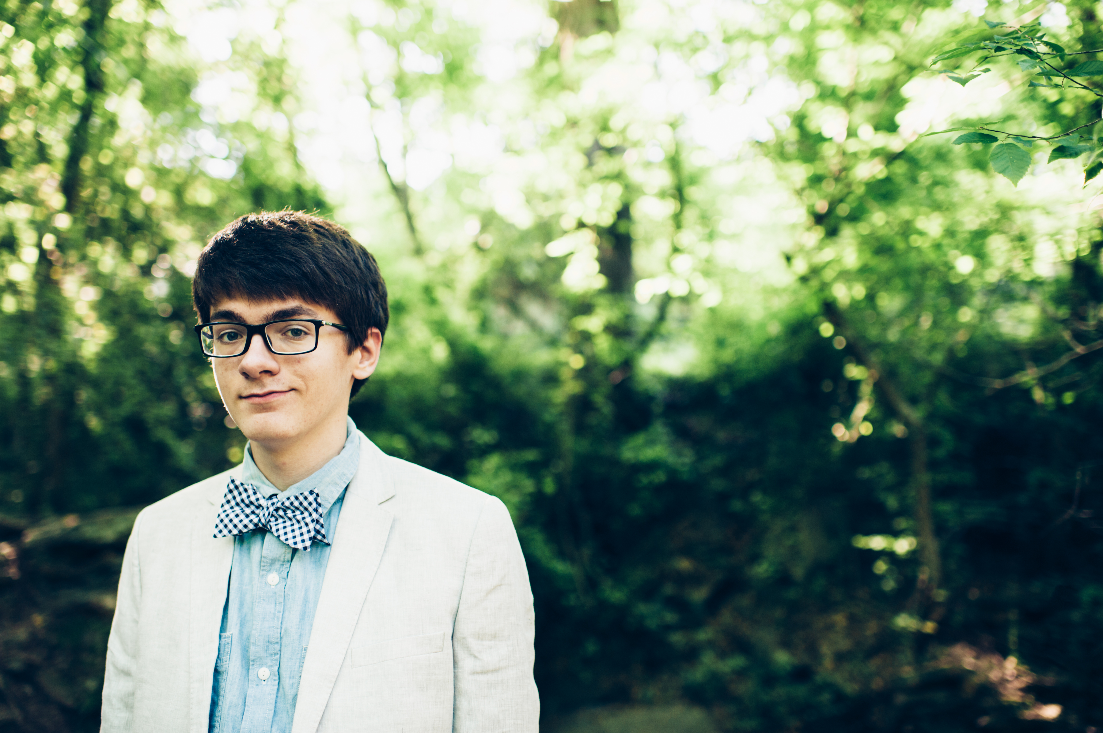 High school senior boy wearing glasses and bow tie smiling slightly in the woods at Sope Creek