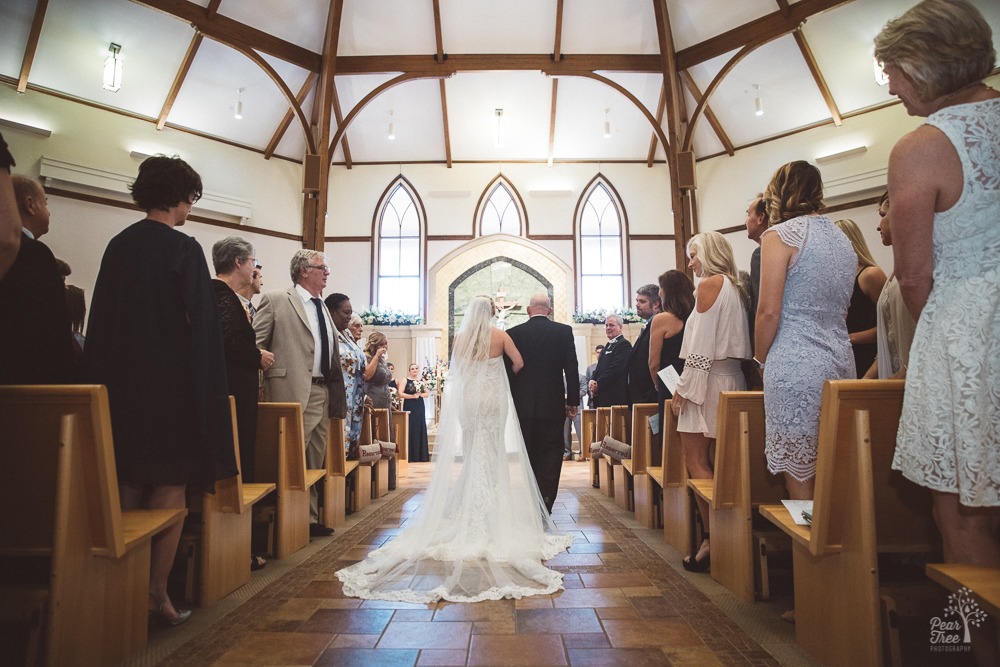 Father walking bride down aisle of St. Ann's Catholic Church to her groom while mom wipes away tears.