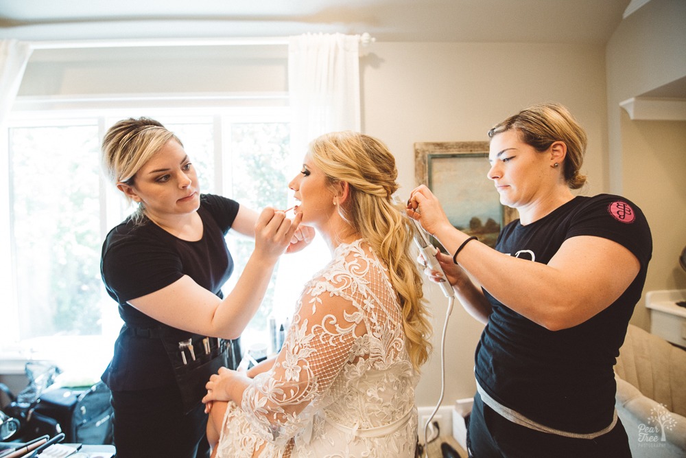 Two assistants applying lip gloss and curls to a beautiful bride in front of window.