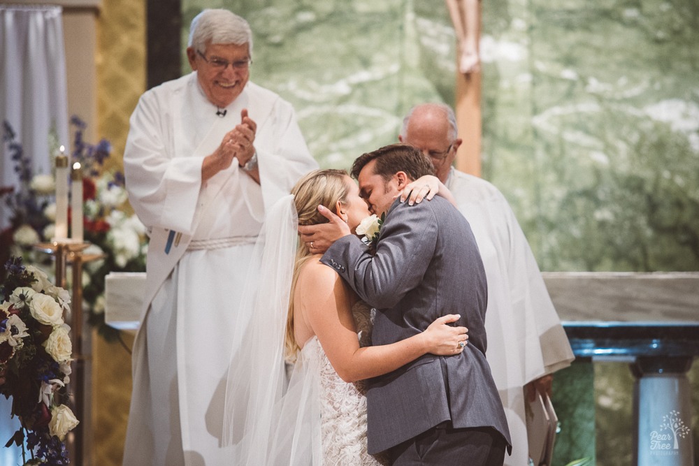 Bride + groom kissing as happy priest looks on and clasps his hands.