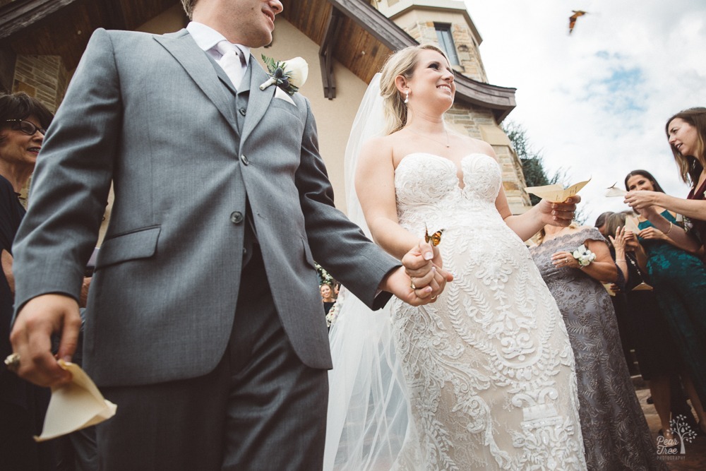 A butterfly lands on bride + grooms hands as they walk out of St. Ann's Catholic Church while other butterflies fly around them.