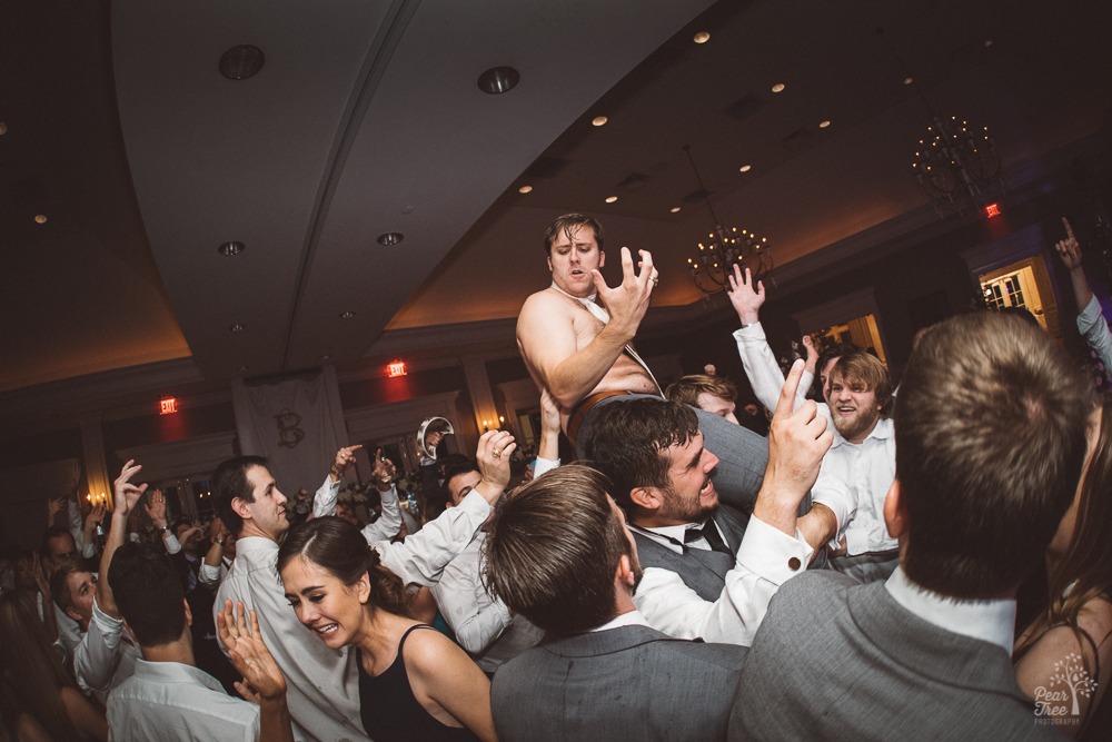 Shirtless groom on shoulders of brother and groomsmen in wedding reception.