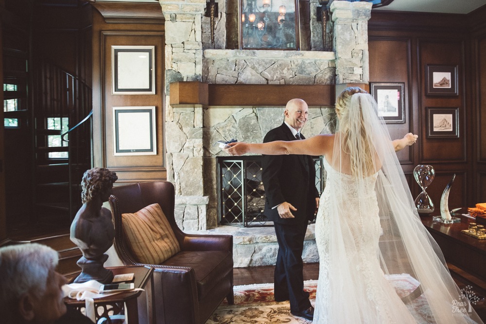 Father seeing daughter with outstretched arms for first time in her wedding gown in a study with a spiral staircase.