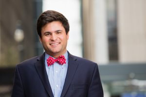 Professional headshot of a smiling man in suit and red bow tie in the middle of an Atlanta city scape