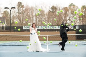 Bride + Groom on tennis court laughing as dozens of tennis balls come at them