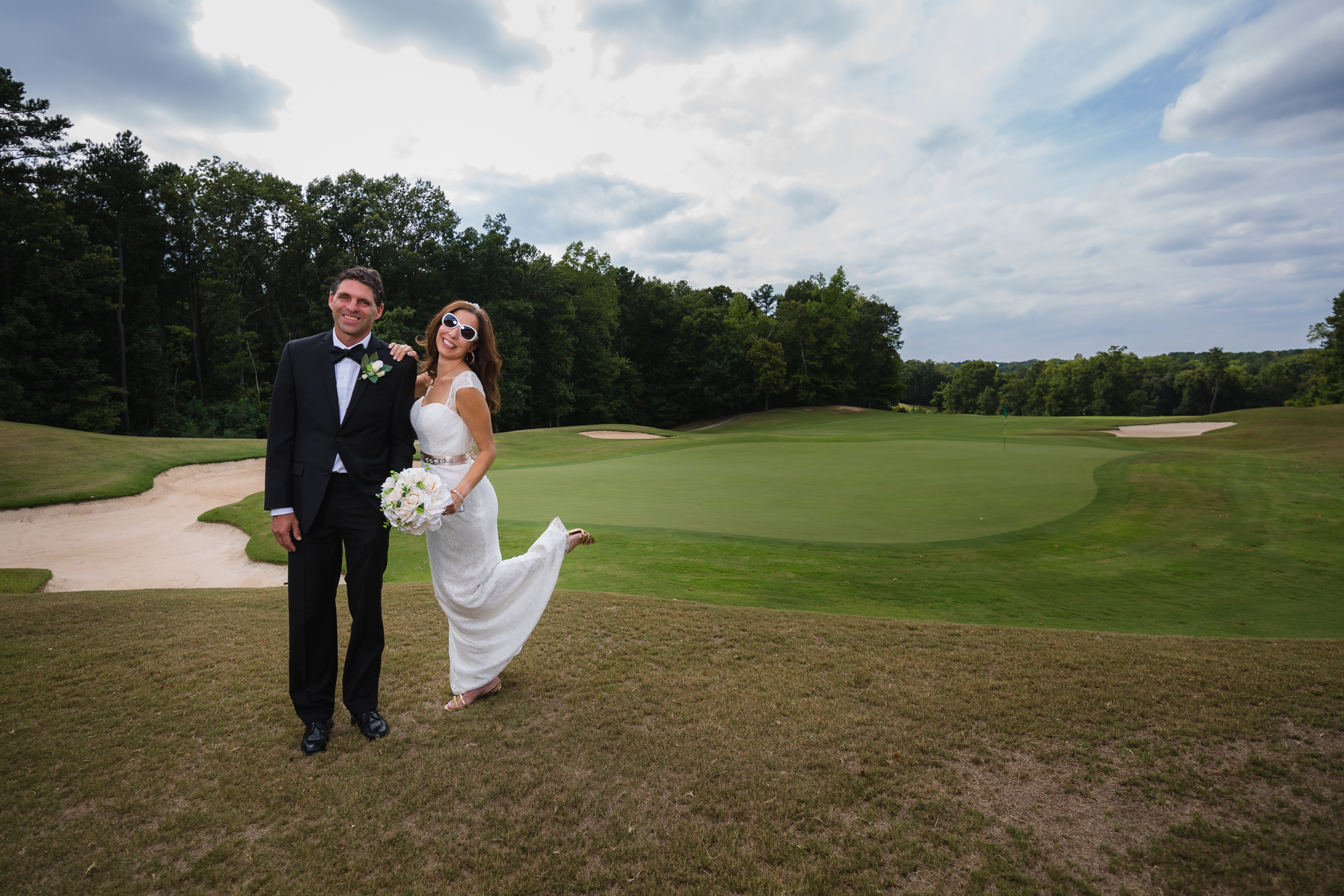 Bride kicking her heel up and smiling in sunglasses while standing next to her husband in front of Stone Mountain golf course