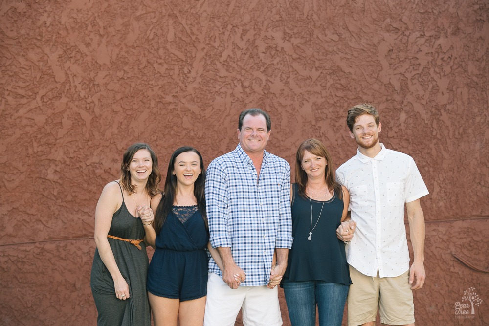 Happy family holding hands and laughing in front of brick red wall with teenage and twenty-something kids