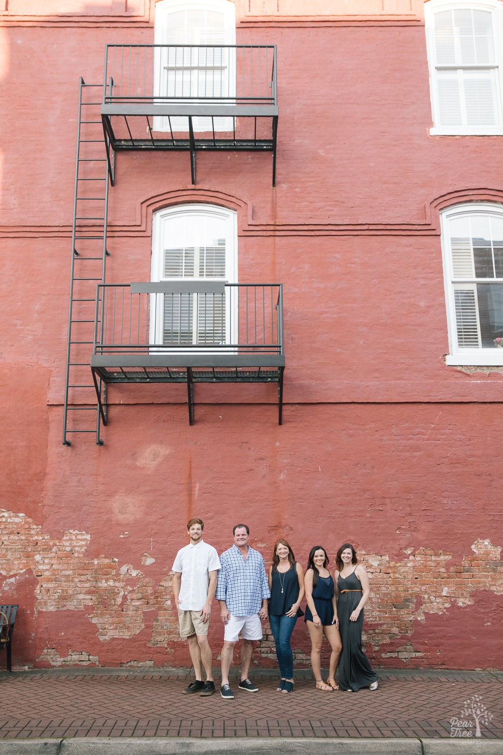 Happy family of two parents and three grown children standing together in downtown Canton under a fire escape in front of an old brick wall.