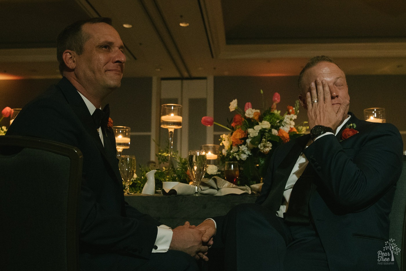 Grooms holding hands and one wiping away tears during wedding toasts.