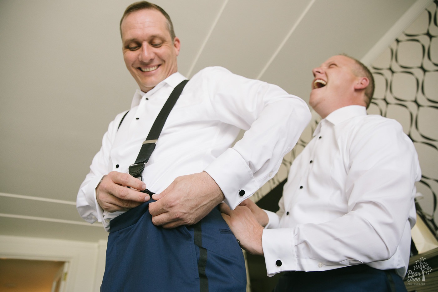 Two grooms laughing and helping fasten suspenders.