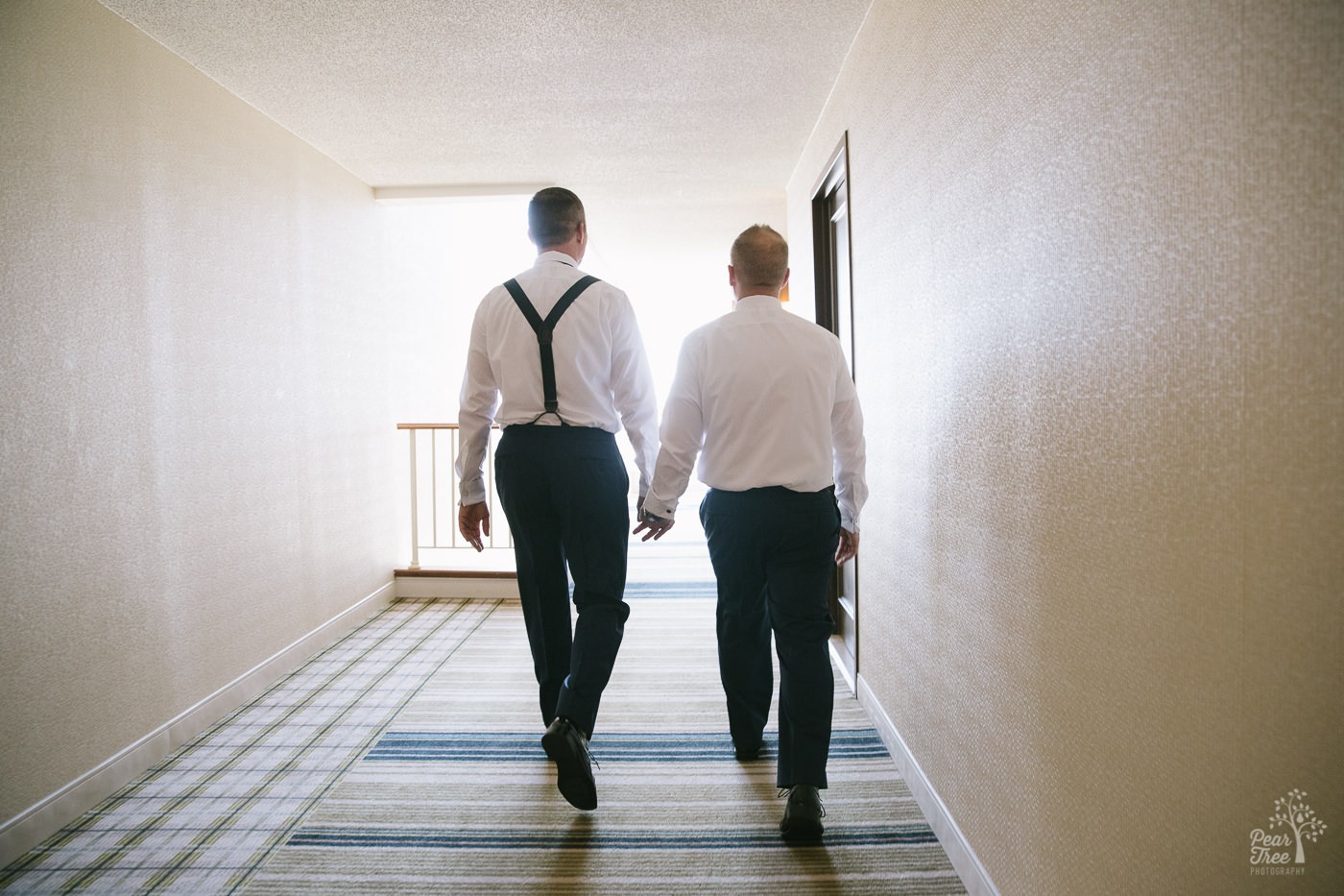 Two grooms holding hands and walking down Renaissance Concourse Atlanta hallway towards the light.