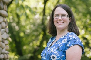 Brunette woman in glasses and blue shirt smiling in front of trees