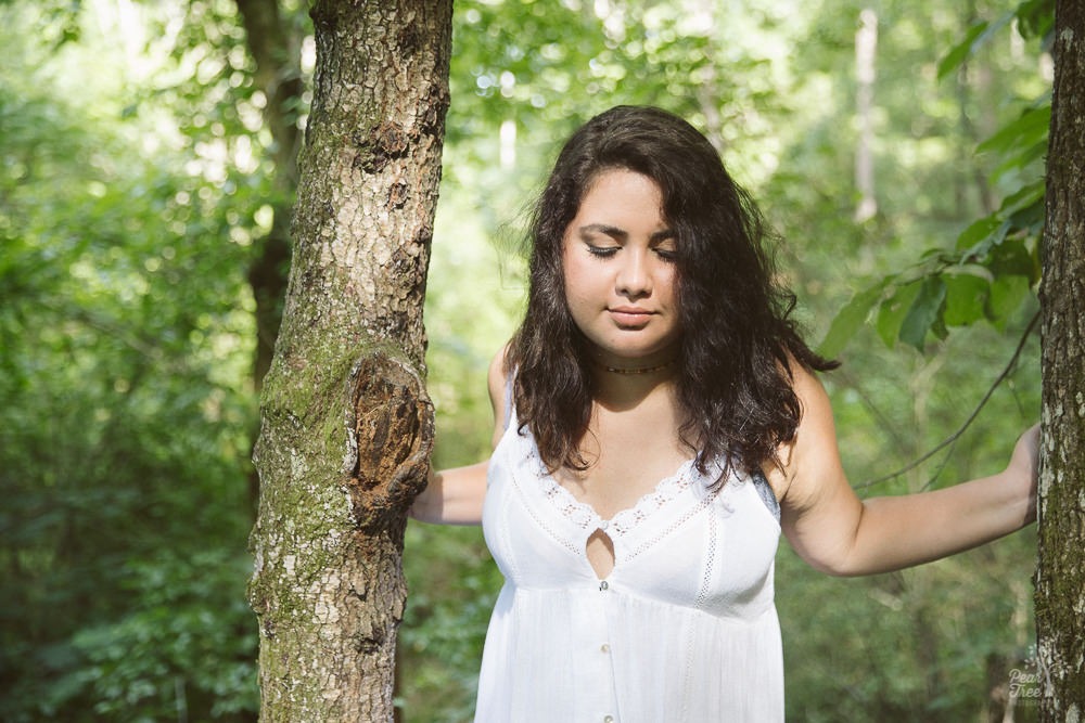 Gorgeous brunette teenage girl standing in woods in white sundress and reflecting on her thoughts.