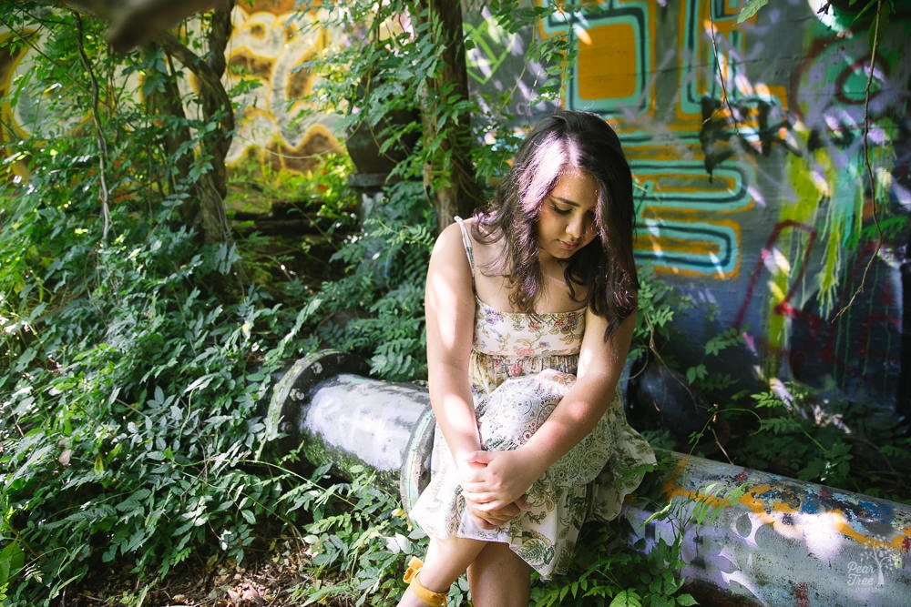 High school girl sitting on Decatur Water Works pipeline surrounded by overgrown foliage and graffiti in the background.