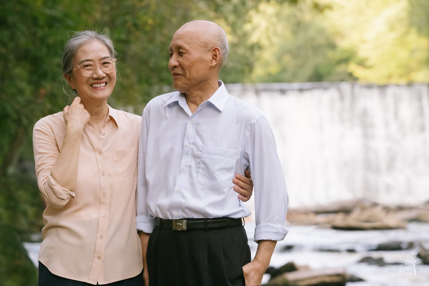 Smiling Chinese woman holding her husband around the waist in front of waterfall at Roswell Mill.