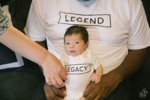 Grandma's hand is straightening out newborn's LEGACY onesie while being held by Dad in LEGEND t-shirt