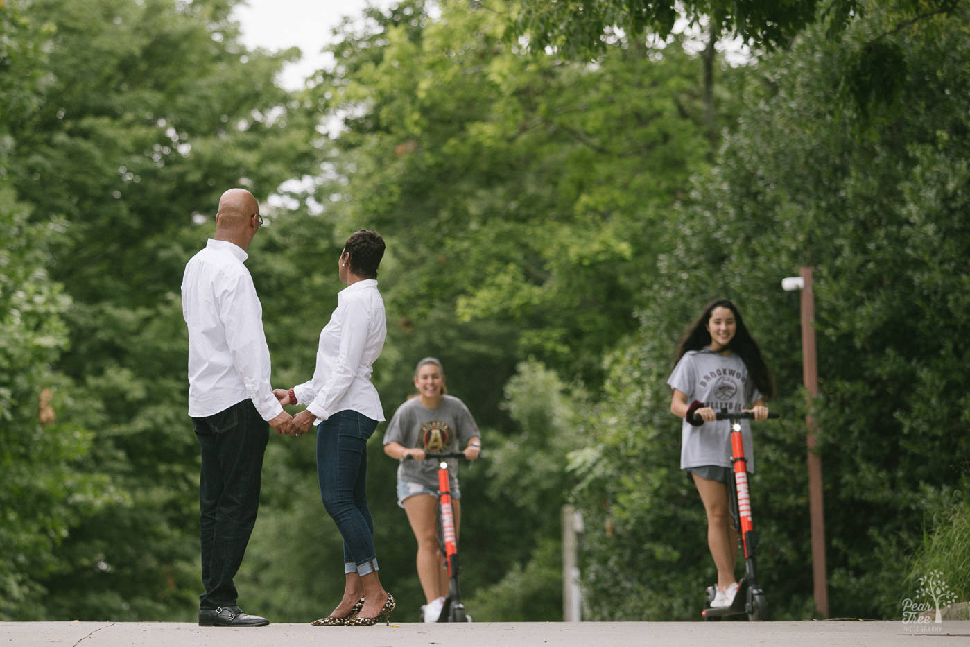 Engaged couple holding hands in Piedmont Park and being photobombed by girls on scooters