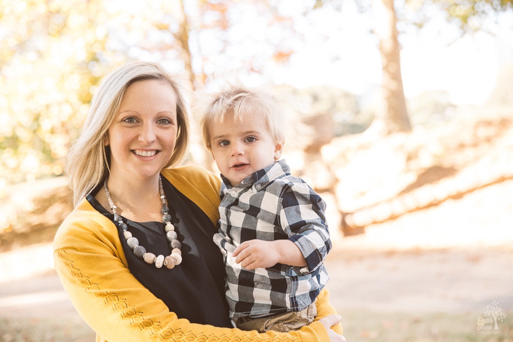 Happy mom holding her cute little toddler boy in front of Piedmont Park fall leaves and trees.