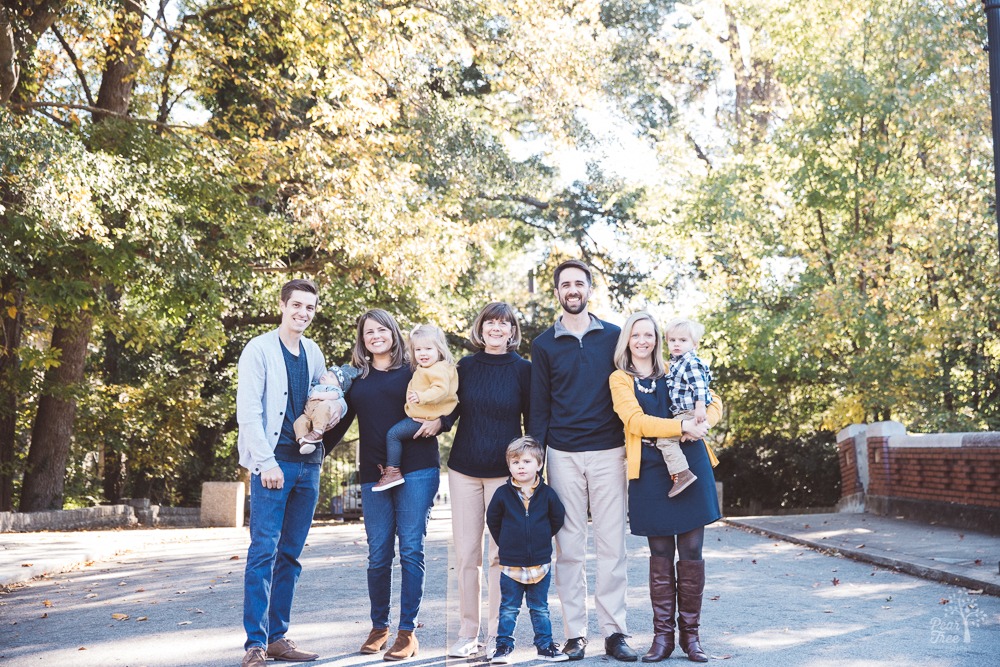 Two families and three generations with Grandma standing in the middle on Piedmont Park bridge in front of fall leaves and trees.