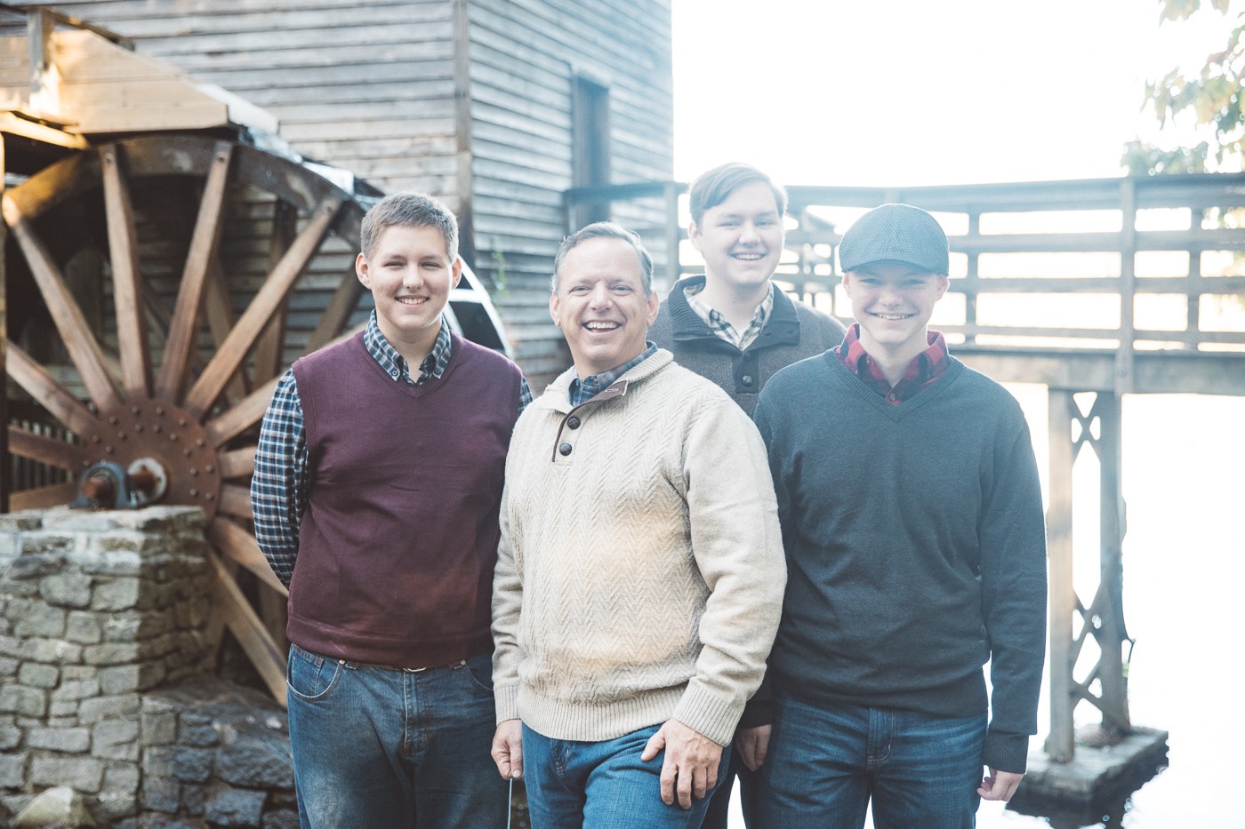 Dad with three teenage sons laughing in front of Stone Mountain grist mill wheel.