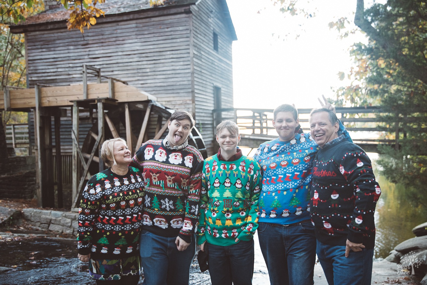 Family of five with three teenage sons wearing ugly Christmas sweaters in front of Stone Mountain old grist mill.