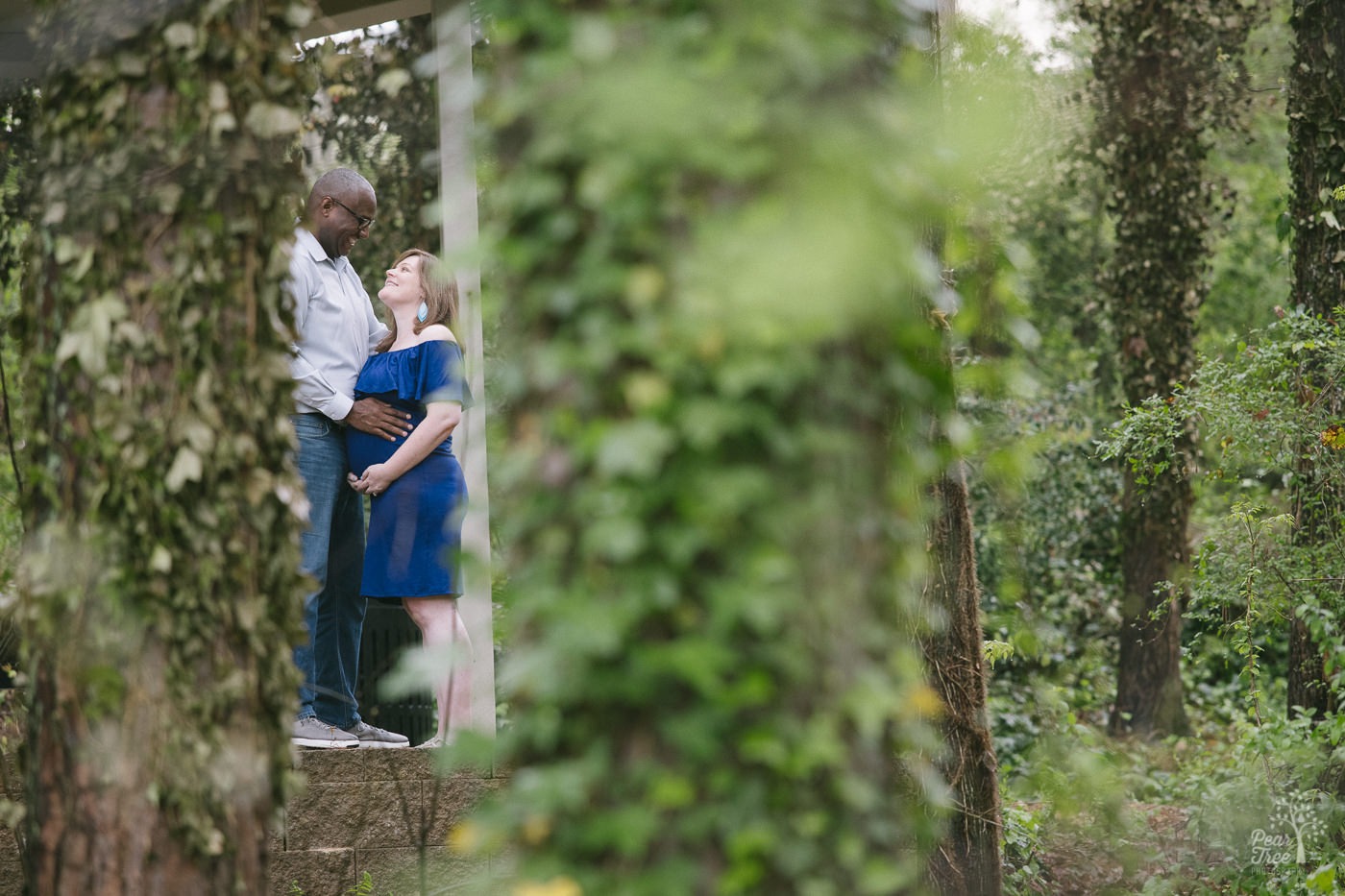 Husband and pregnant wife standing close smiling and holding her belly in the middle of a forest.