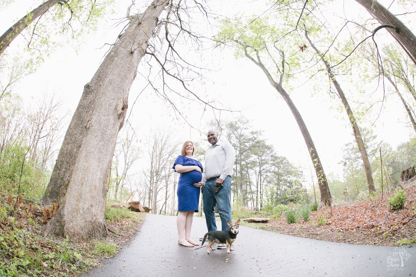 Pregnant mom in blue dress holding her belly while standing next to her husband on a trail with their little dog.