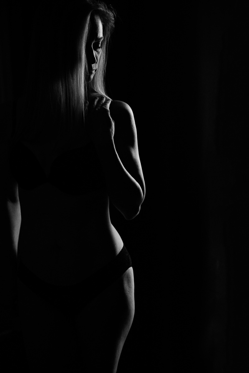 Silhouette of a woman standing in a sliver of light with her hand on her shoulder and underwear band visible over her hip