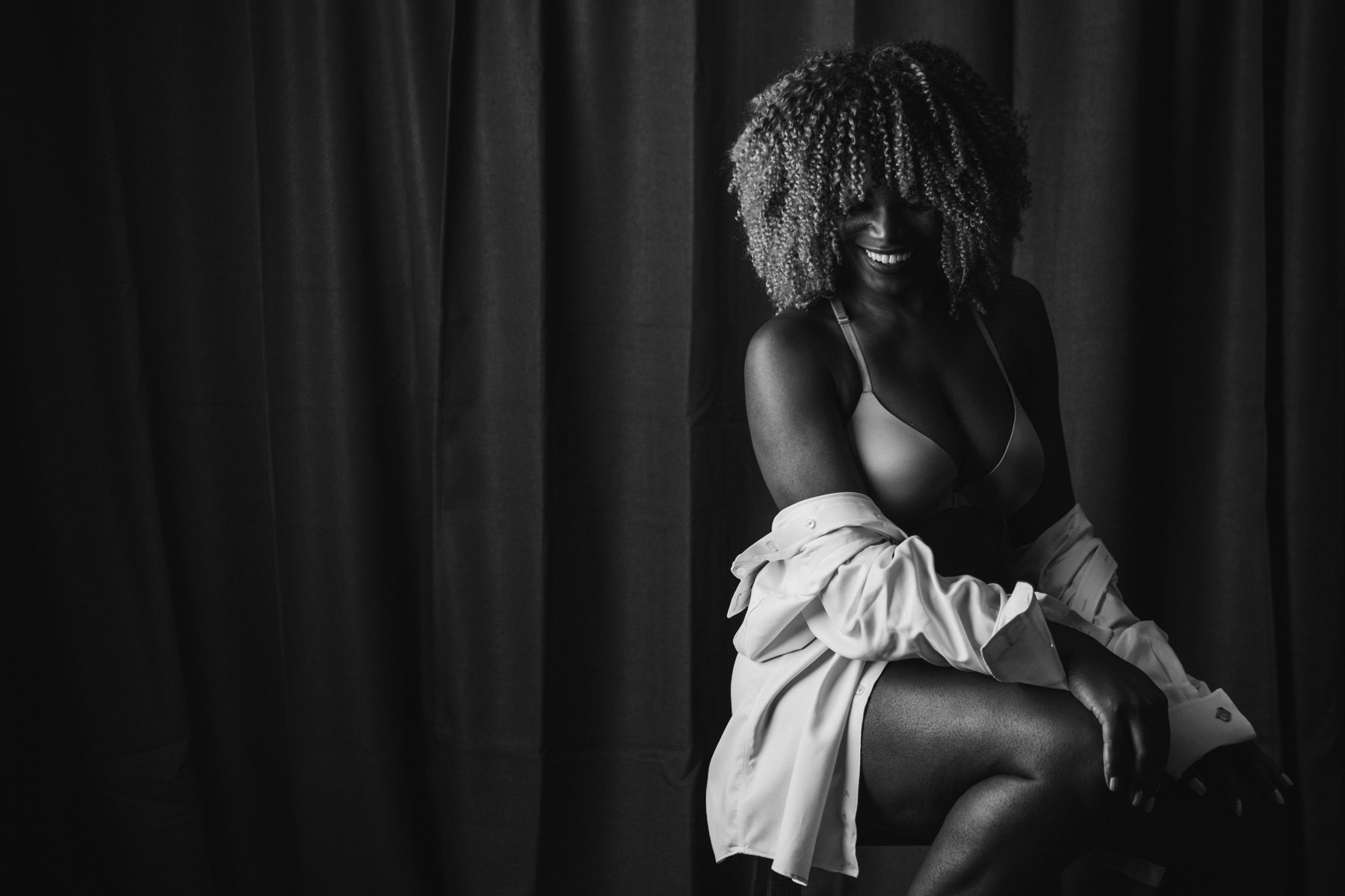 A confidently, gorgeous black woman sitting on a stool in her lingerie and man's white dress shirt while laughing during a boudoir session