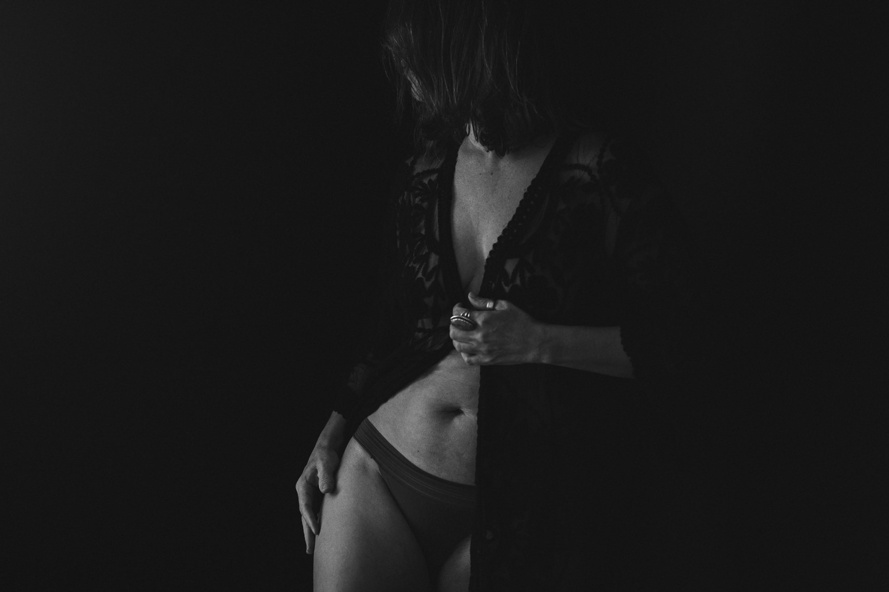 Black and white boudoir photograph of a faceless woman in a lace robe held together under her breasts with one hand and other hand on her hip over underwear