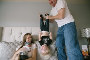 Mom holding newborn son on bed while Dad stands and holds their toddler upside down