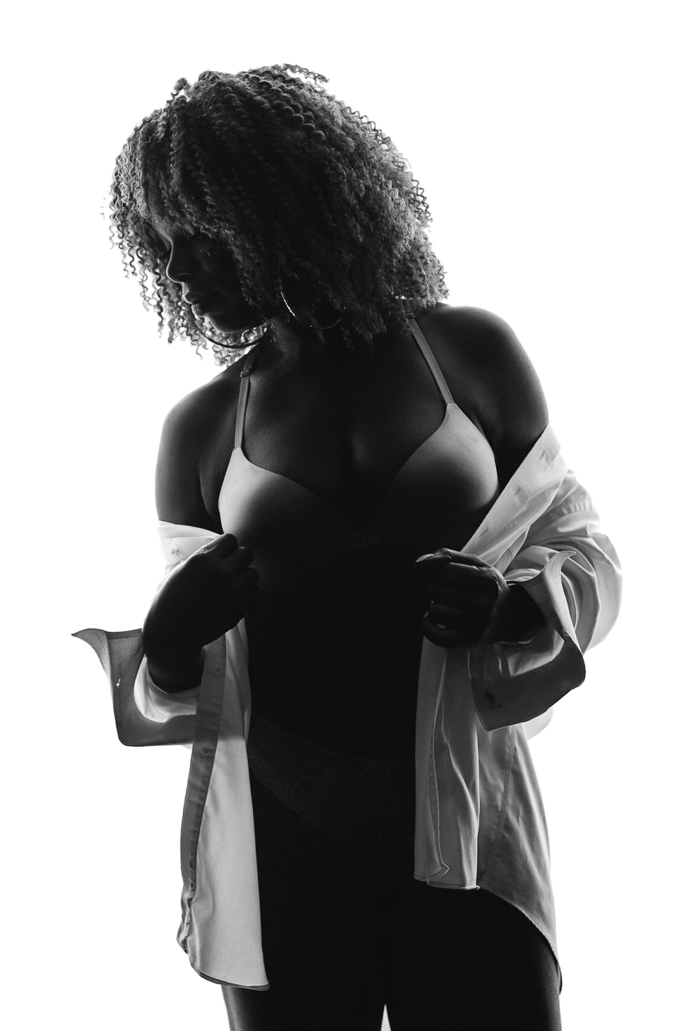 Gorgeous African American woman with amazing hair standing in partial silhouette while pulling boyfriend's white work shirt around her body and off her shoulders for boudoir photographs