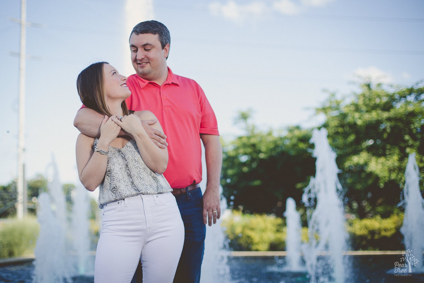 Handsome husband wrapping his arm around his wife's chest and arm to hold her close in front of fountains as they smile at each other