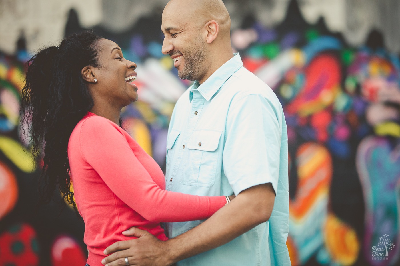 African American couple holding each other close and laughing hard in front of a colorful mural for anniversary photos