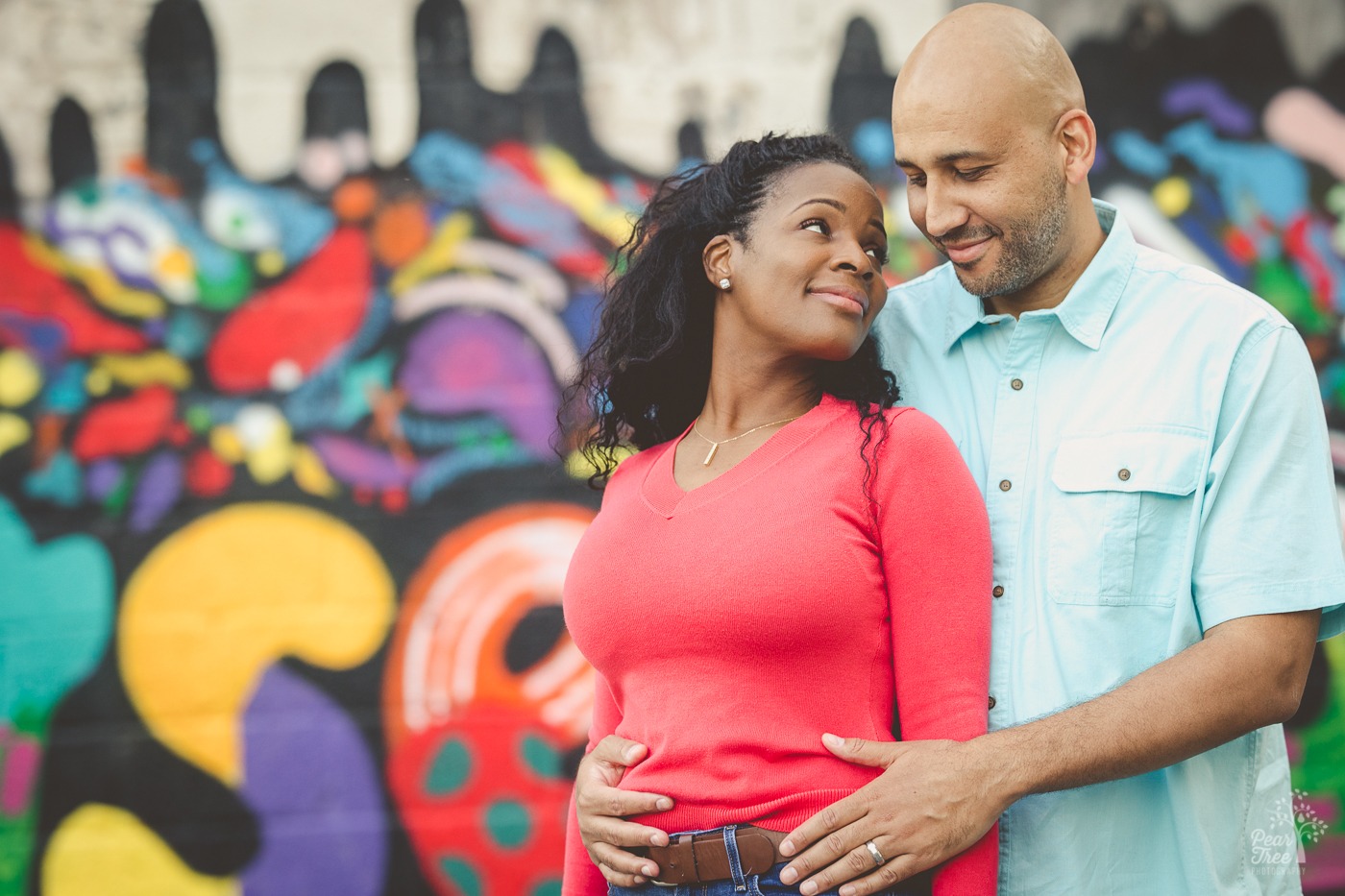 African American wife smiling up at her husband while he holds her close with his eyes closed while standing in front of a colorful mural