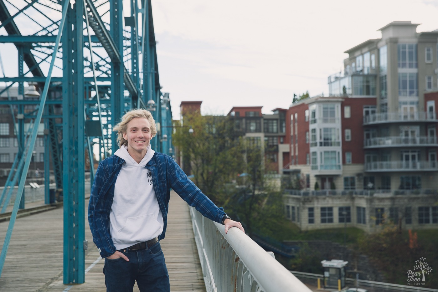 Blond high school senior boy wearing white hoodie, blue flannel shirt, and jeans standing smiling on blue Chattanooga bridge