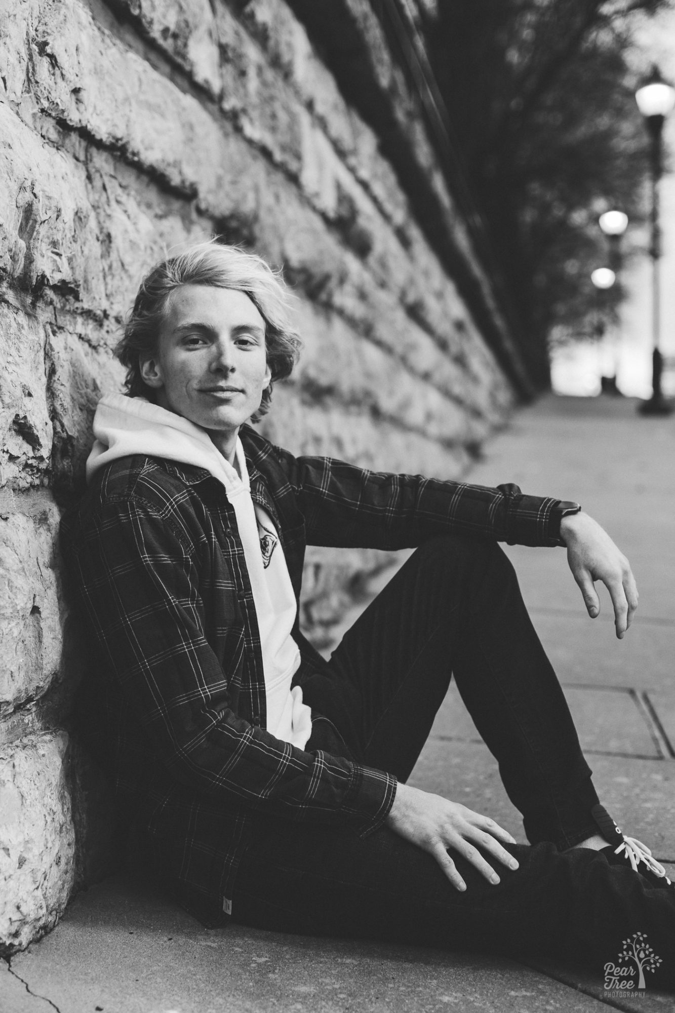 Black and white photo of high school senior boy sitting on sidewalk against a stone wall and his arm resting on his raised knee