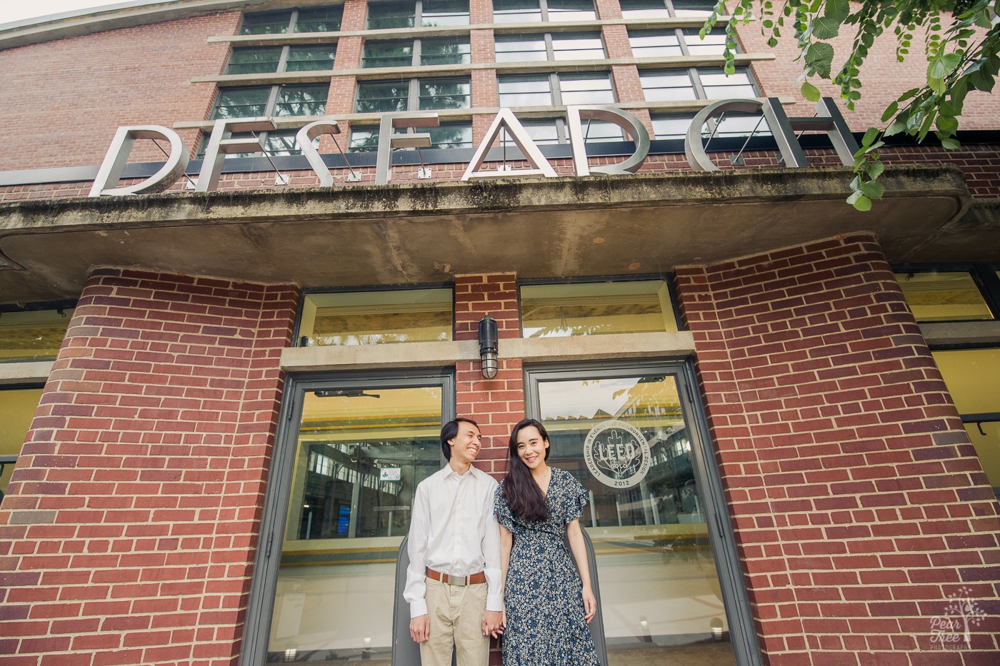 Engaged Asian-American couple holding hands and smiling in front of  Georgia Tech architectural graduate studies building