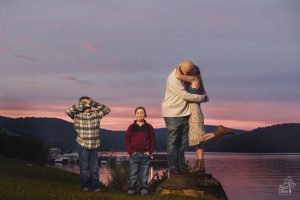 Husband and wife hugging and kissing at sunset while their sons look creeped out
