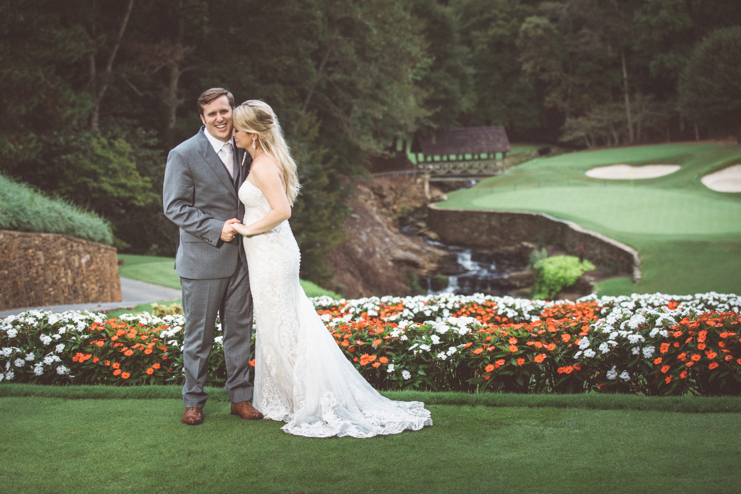 Atlanta wedding photograph of laughing bride and groom holding hands and embracing closely at top of Atlanta Country Club golf course on their wedding day with beautiful red and white flowers and a little creek behind them