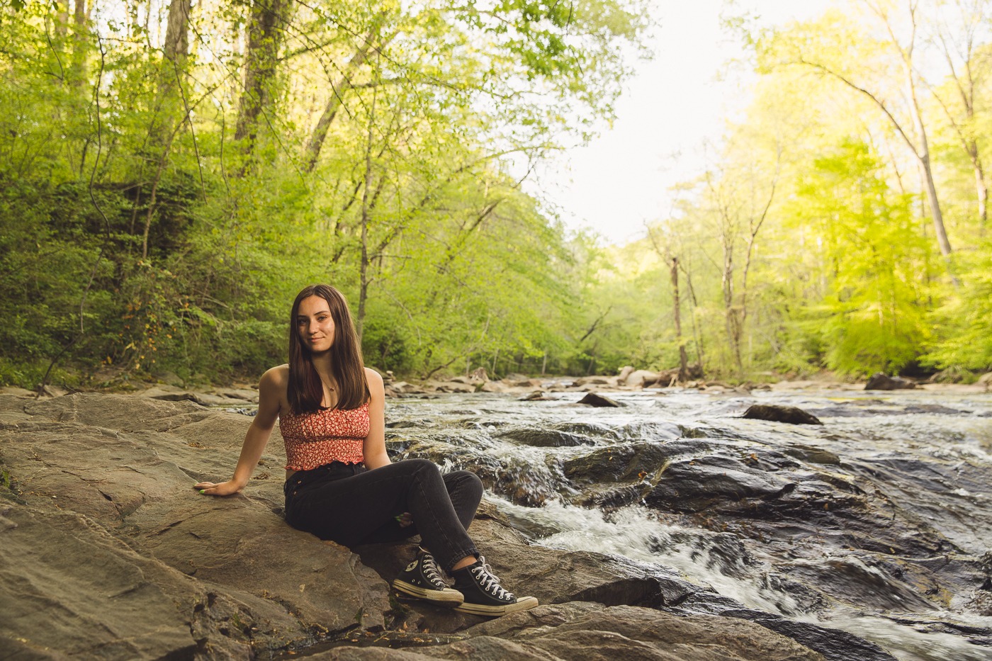 Gorgeous high school senior girl sitting in her black jeans and Converse with the river rushing around her on the shoals