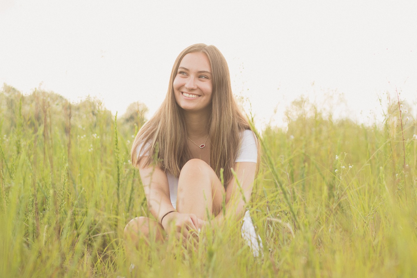 Happy high school senior girl sitting in tall grass hugging her knee and wearing a simple ruby red necklace