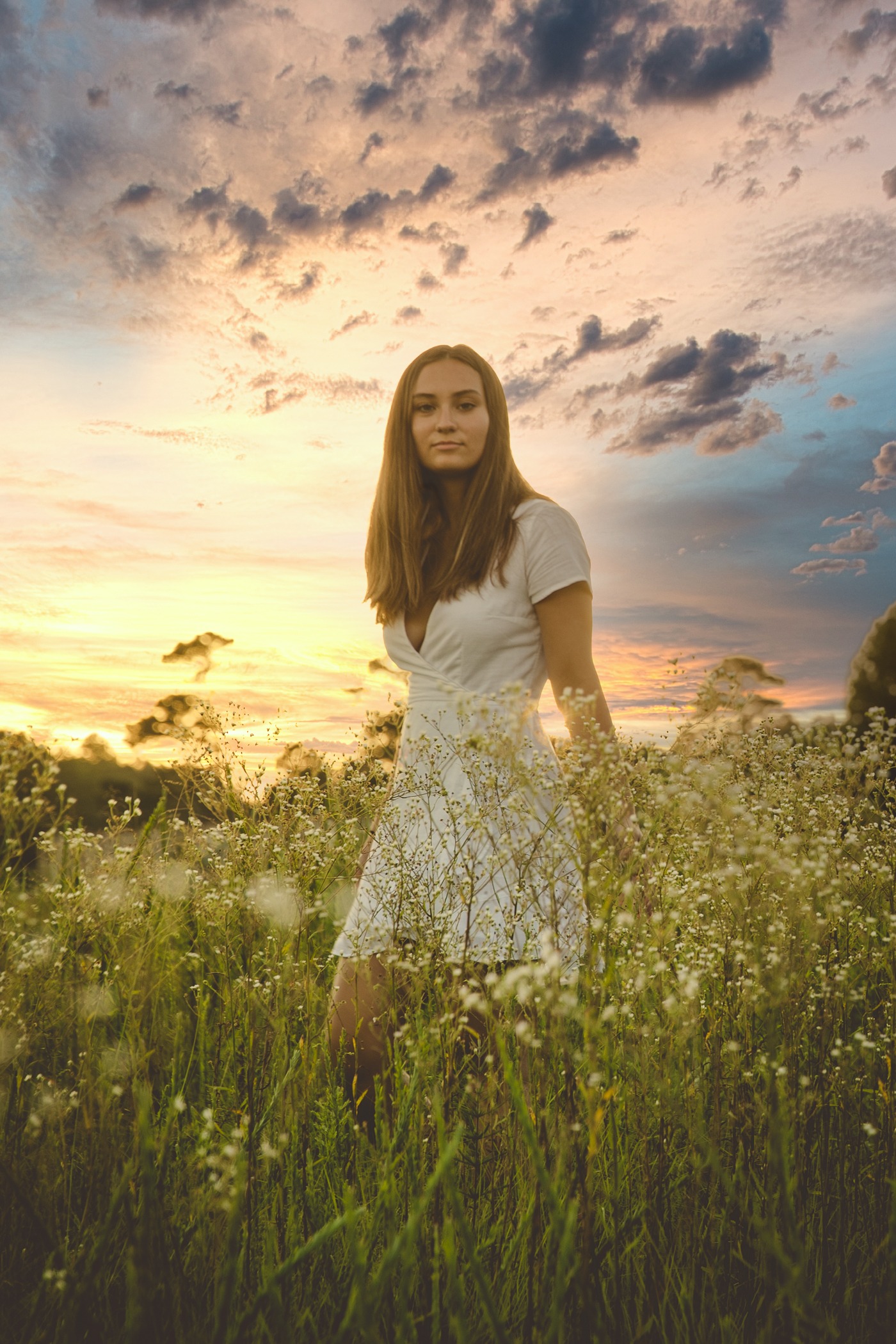 Thoughtful and pretty high school senior girl standing in white wildflowers in front of a dramatic sunset